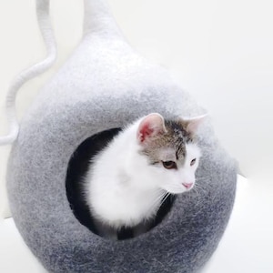 May include: A white and gray felted cat bed with a round opening. A white cat with gray markings is sitting inside the bed.