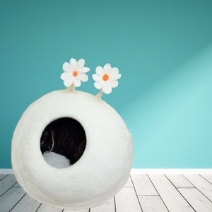 May include: A white felt cat bed shaped like a sphere with two white felt flowers on top. The bed has a round opening for the cat to enter. A cat is sleeping inside the bed.