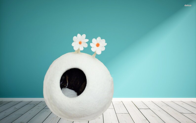 May include: A white felt cat bed shaped like a sphere with two white felt flowers on top. The bed has a round opening for the cat to enter. A cat is sleeping inside the bed.