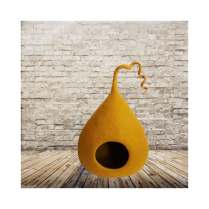 May include: A mustard yellow felt cat bed shaped like a teardrop with a curled felt tail. The bed is sitting on a wooden floor.