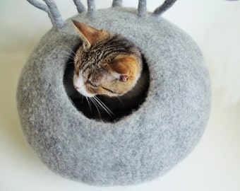 May include: A gray felt cat bed with a round opening. A tabby cat is peeking out of the opening.