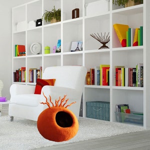 Peut inclure: Lit de chat en feutre orange avec un trou d'entr&eacute;e et un design unique de brins de feutre s'&eacute;tendant du haut. Le lit est pos&eacute; sur un tapis blanc dans une pi&egrave;ce avec un fauteuil blanc et une biblioth&egrave;que blanche.