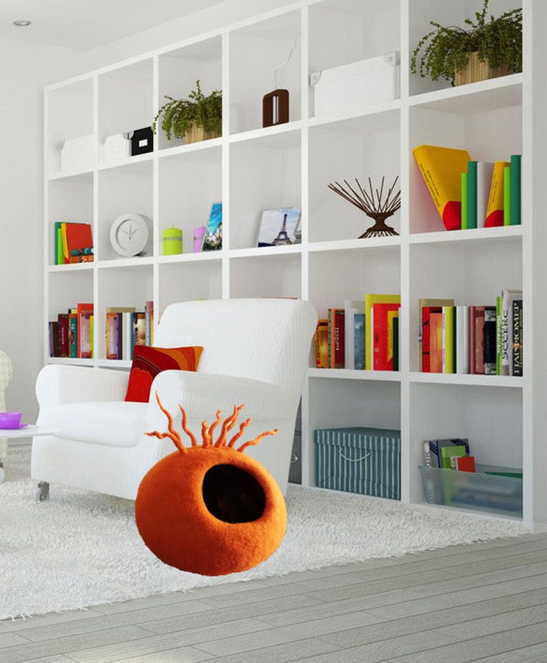 Peut inclure: Lit de chat en feutre orange avec un trou d'entr&eacute;e et un design unique de brins de feutre s'&eacute;tendant du haut. Le lit est pos&eacute; sur un tapis blanc dans une pi&egrave;ce avec un fauteuil blanc et une biblioth&egrave;que blanche.
