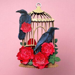 May include: A decorative wooden birdcage featuring two black crows and vibrant red roses. The cage is a warm gold colour and has the text "MYHELLOWOOD" on it. The roses are clustered around the base and inside the cage. The background is pink.