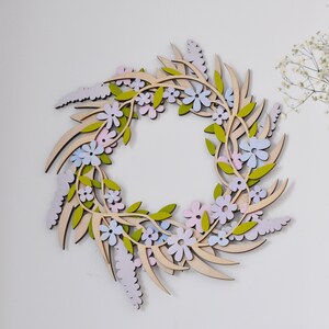 Puede incluir: Una corona de madera decorada con flores de color rosa pastel, azul y verde. La corona est&aacute; hecha de madera y tiene un acabado de madera natural.