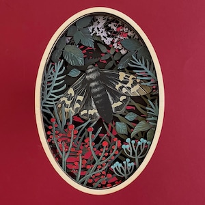 May include: An oval-shaped, layered paper art piece featuring a moth surrounded by foliage. The moth is black with tan and yellow markings. The artwork includes red berries, blue flowers, and green leaves, all set against a deep red background.