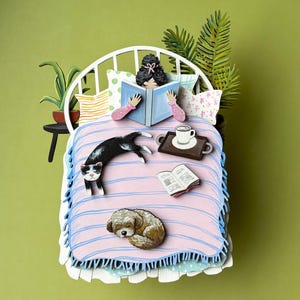 Può includere: Un'illustrazione stravagante di una persona che legge a letto, circondata da un gatto bianco e nero, un cane che dorme, una tazza di caffè e un libro. Il letto ha una coperta rosa a righe blu su sfondo verde.