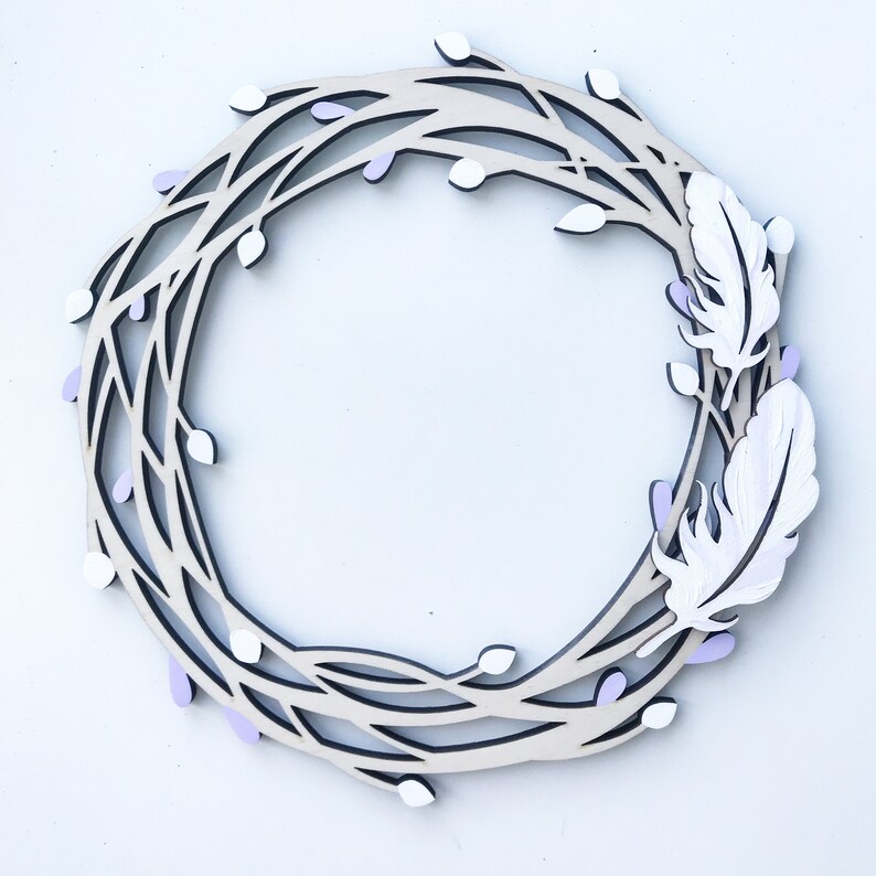 May include: A wooden wreath with a delicate, intricate design featuring branches and small purple flowers. Two white feathers are attached to the wreath, adding a touch of whimsy.