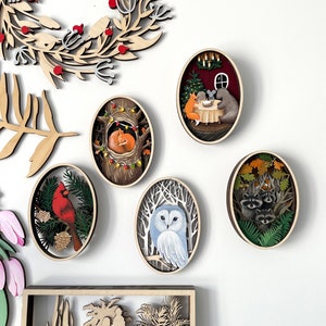 May include: Five wooden oval ornaments with intricate cutouts and painted scenes. The ornaments feature a cardinal on a branch, a fox sleeping in a tree, an owl in a snowy forest, raccoons in a forest, and a fox, bear, and mouse at a table with a Christmas tree.
