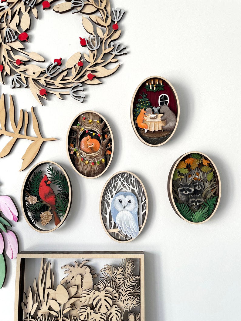 May include: Five wooden oval ornaments with intricate cutouts and painted scenes. The ornaments feature a cardinal on a branch, a fox sleeping in a tree, an owl in a snowy forest, raccoons in a forest, and a fox, bear, and mouse at a table with a Christmas tree.