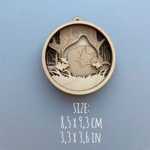 May include: A wooden ornament featuring a laser-cut design of a fox sleeping under a tree. The ornament is 8.5 cm by 9.3 cm.