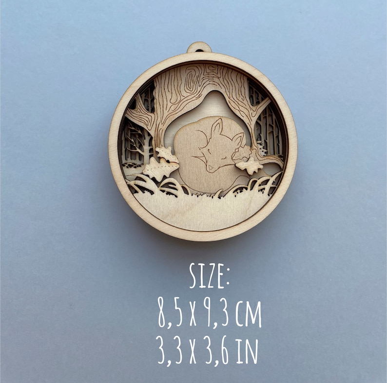 May include: A wooden ornament featuring a laser-cut design of a fox sleeping under a tree. The ornament is 8.5 cm by 9.3 cm.