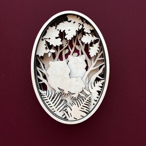 May include: A white wooden oval-shaped wall decor with a laser-cut design of two foxes in a forest setting. The foxes are surrounded by trees, leaves, and ferns.