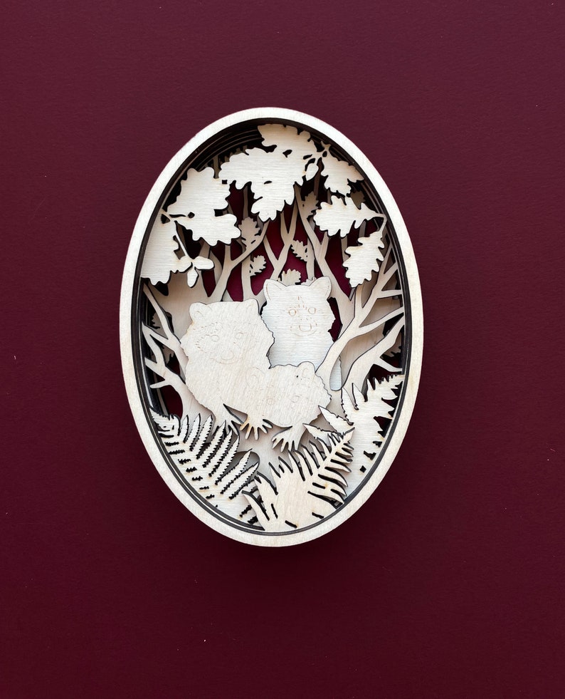May include: A white wooden oval-shaped wall decor with a laser-cut design of two foxes in a forest setting. The foxes are surrounded by trees, leaves, and ferns.