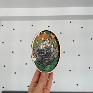 May include: A wooden oval wall decor with a 3D layered design of raccoons in a forest setting. The raccoons are depicted in a tree with leaves and ferns. The background is a light wood color.