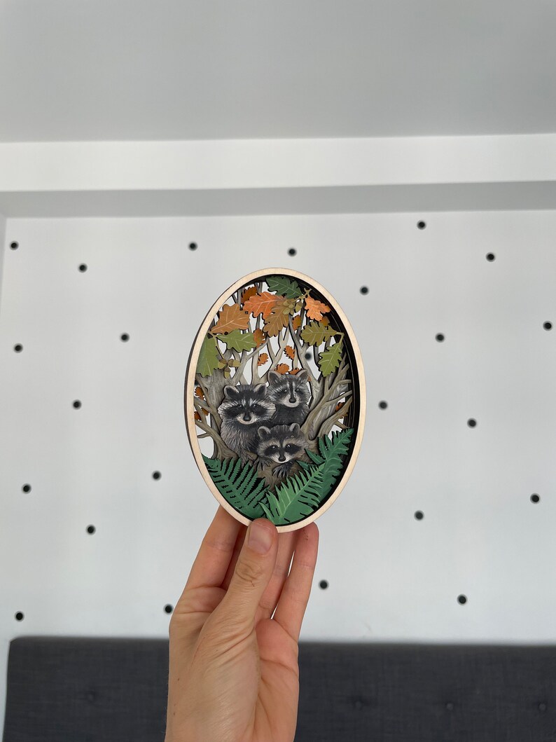 May include: A wooden oval wall decor with a 3D layered design of raccoons in a forest setting. The raccoons are depicted in a tree with leaves and ferns. The background is a light wood color.
