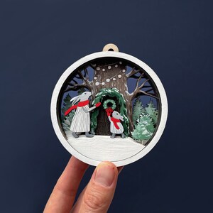 May include: A white wooden ornament depicting two rabbits decorating a tree with a wreath and string lights. The ornament is painted with details of a snowy forest scene.
