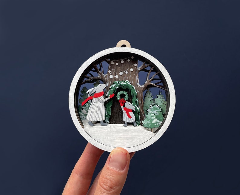 May include: A white wooden ornament depicting two rabbits decorating a tree with a wreath and string lights. The ornament is painted with details of a snowy forest scene.