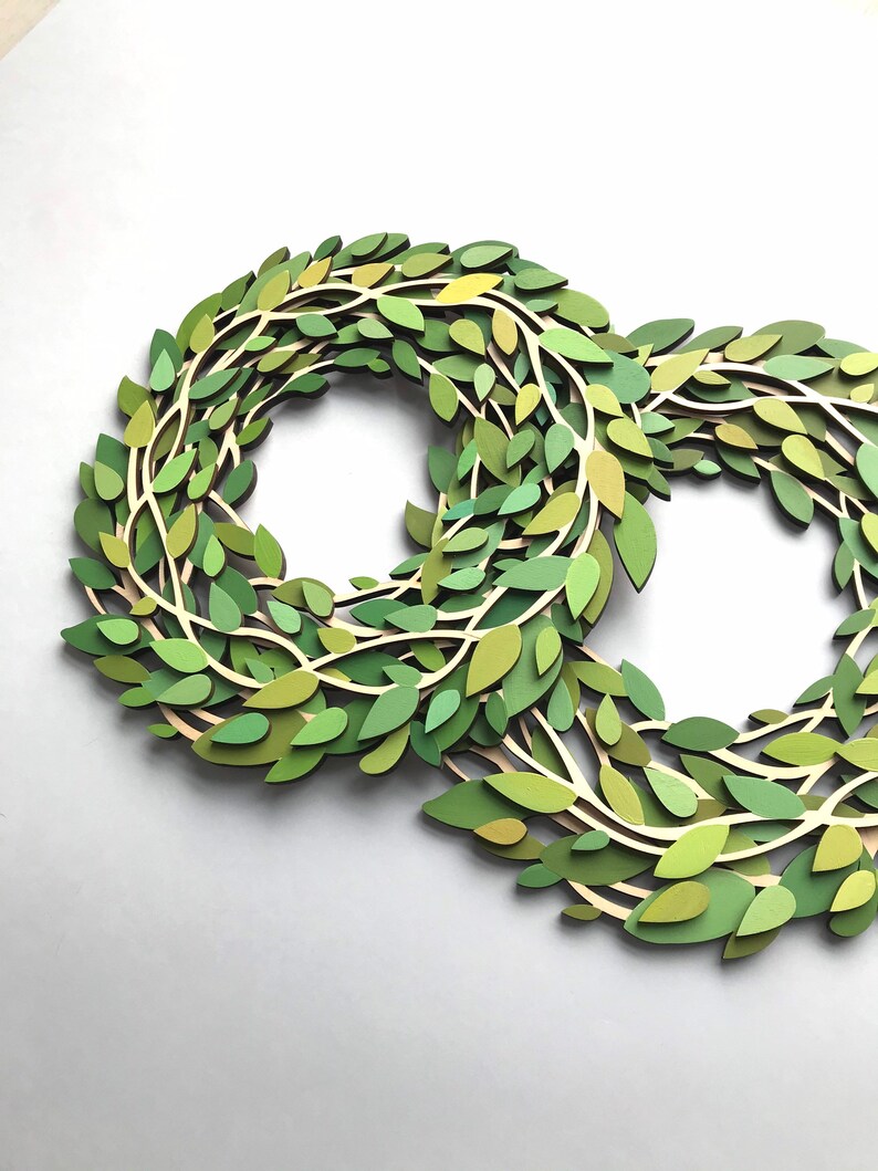 May include: Two green paper wreaths made of layered leaves, with a light brown background.
