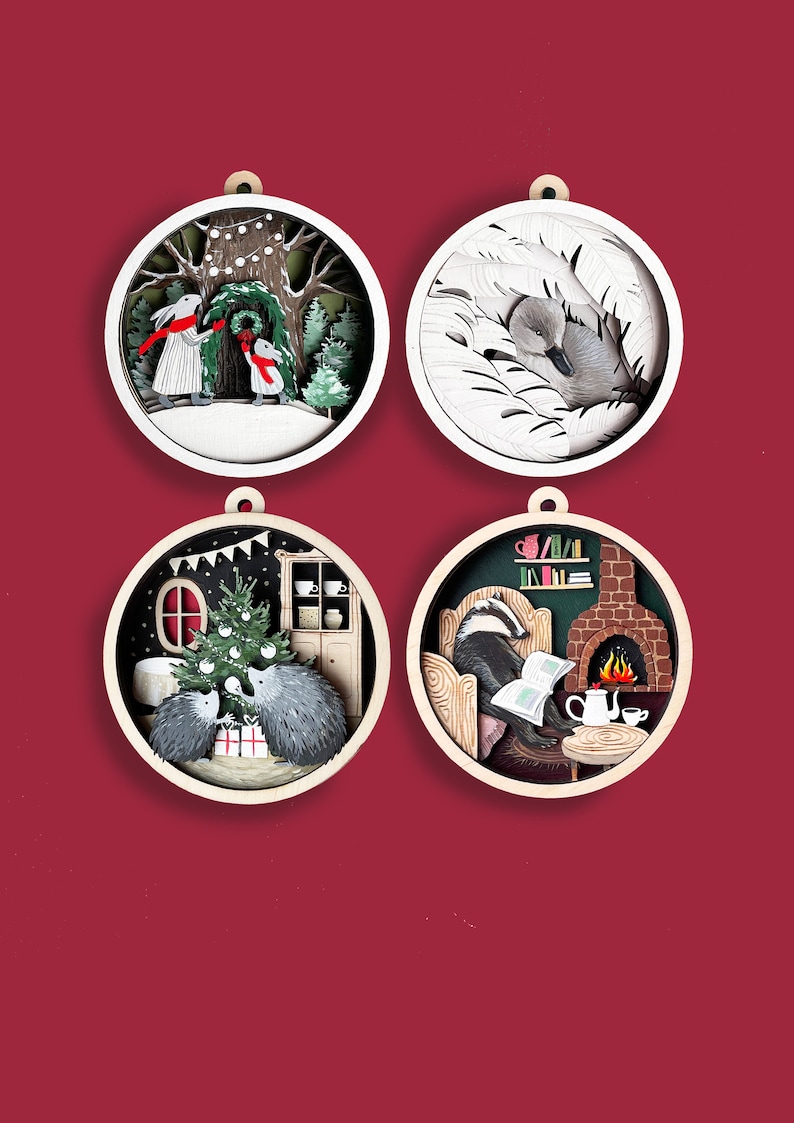 May include: Four round wooden ornaments with paper cut-out designs. The first ornament features two rabbits in a snowy forest. The second ornament features a swan in a nest of feathers. The third ornament features two hedgehogs decorating a Christmas tree. The fourth ornament features a badger reading a book by a fireplace.