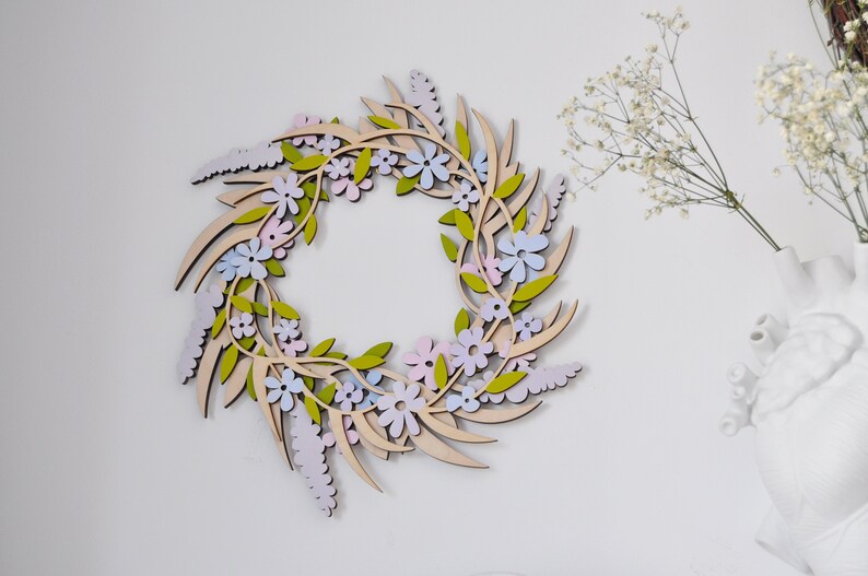 Puede incluir: Una corona de madera decorada con flores de color rosa pastel, azul y verde. La corona est&aacute; hecha de madera y tiene un acabado de madera natural.