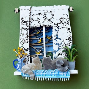 Op de afbeelding: Papierkunstscène van een raam met witte kanten gordijnen, vogels en twee katten. Een blauw-wit gestreepte vaas met gele bloemen en een potplant staan op de vensterbank. De katten zijn grijs en bruin, rustend op een blauw geruit kussen.