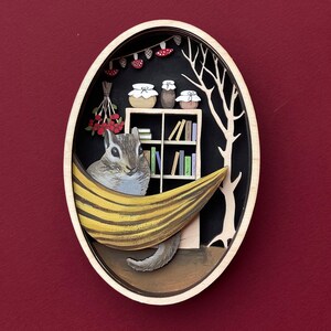 May include: A chipmunk in a yellow hammock, resting in a wooden oval frame. The frame features a black background with a bookshelf, a tree, and red berries.