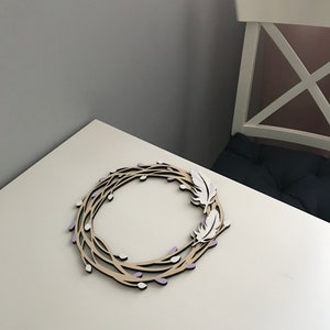 May include: A wooden wreath with a natural brown finish, decorated with white feathers and purple accents. The wreath is made of branches with small buds.