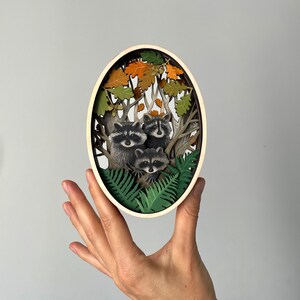 May include: A wooden oval-shaped wall art piece with a 3D paper cut-out design of three raccoons in a forest setting. The raccoons are surrounded by green ferns and brown oak leaves.