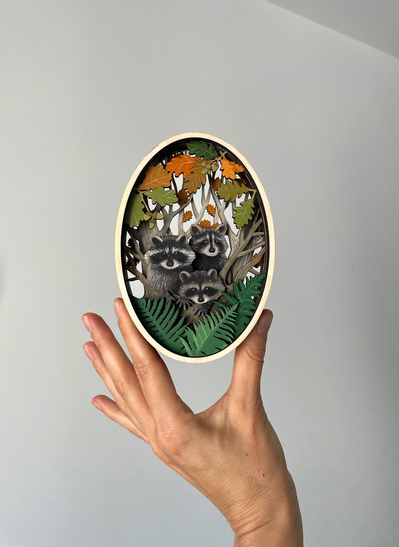 May include: A wooden oval-shaped wall art piece with a 3D paper cut-out design of three raccoons in a forest setting. The raccoons are surrounded by green ferns and brown oak leaves.