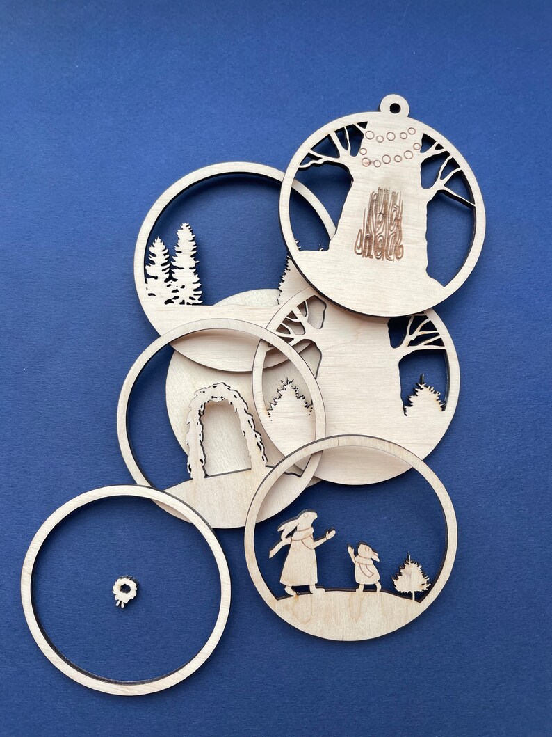May include: Six wooden ornaments with various winter-themed designs, including a tree, a wreath, an archway, and a pair of rabbits. The ornaments are all cut out of wood and have a natural finish.