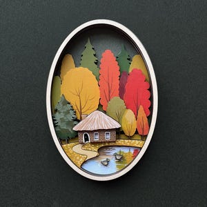 Op de afbeelding: Een ovaal houten frame met een 3D-papieren kunstscène van een klein huis in een bos. De bomen zijn in verschillende tinten groen, geel, oranje en rood. Er is een klein vijvertje voor het huis met twee eenden die erin zwemmen.