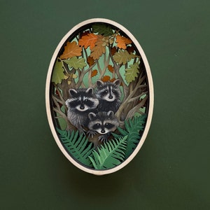 May include: A wooden oval-shaped wall decor with a 3D paper cut-out design of three raccoons in a forest setting. The raccoons are surrounded by leaves and branches, and the background is a dark green.