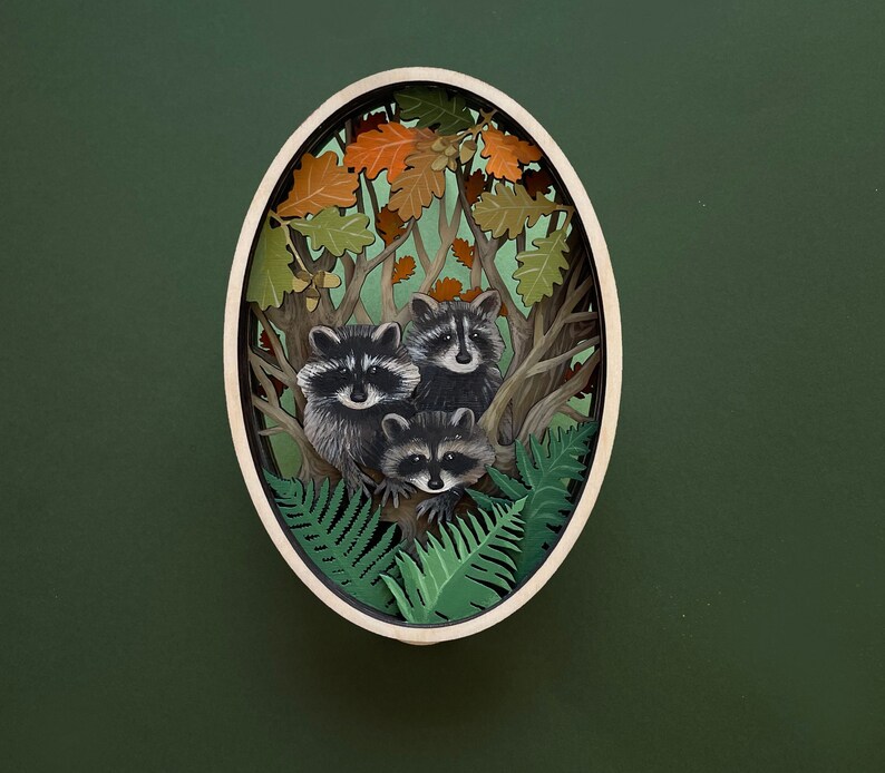 May include: A wooden oval-shaped wall decor with a 3D paper cut-out design of three raccoons in a forest setting. The raccoons are surrounded by leaves and branches, and the background is a dark green.