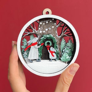 May include: White wooden ornament with a winter scene. Two white rabbits with red scarves are decorating a tree with a wreath. The tree is surrounded by snow-covered pine trees and a white background.