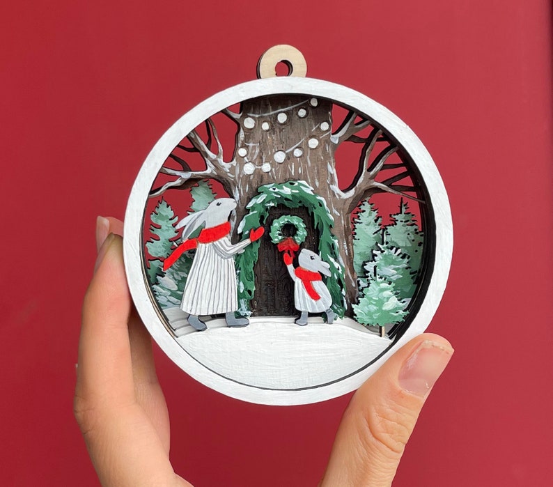 May include: White wooden ornament with a winter scene. Two white rabbits with red scarves are decorating a tree with a wreath. The tree is surrounded by snow-covered pine trees and a white background.