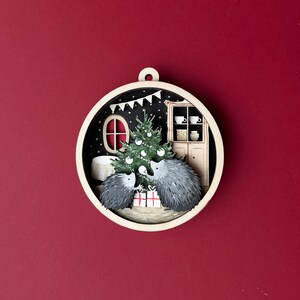 May include: A wooden Christmas ornament featuring two hedgehogs under a decorated Christmas tree in a cozy room with a fireplace and a cabinet. The ornament is painted in a whimsical style with a black background and white accents.