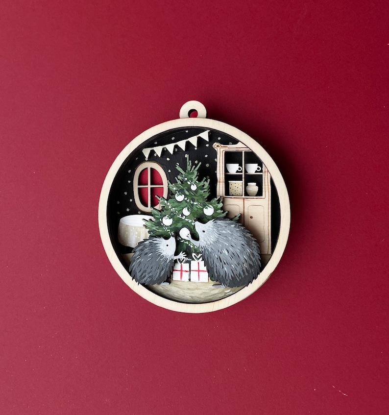 May include: A wooden Christmas ornament featuring two hedgehogs under a decorated Christmas tree in a cozy room with a fireplace and a cabinet. The ornament is painted in a whimsical style with a black background and white accents.