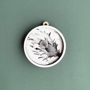 May include: A white wooden ornament with a cutout design of a bird nestled in leaves. The bird is gray and white and has its head tucked under its wing.