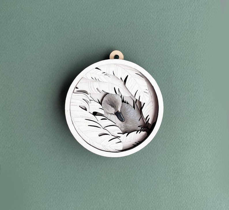 May include: A white wooden ornament with a cutout design of a bird nestled in leaves. The bird is gray and white and has its head tucked under its wing.