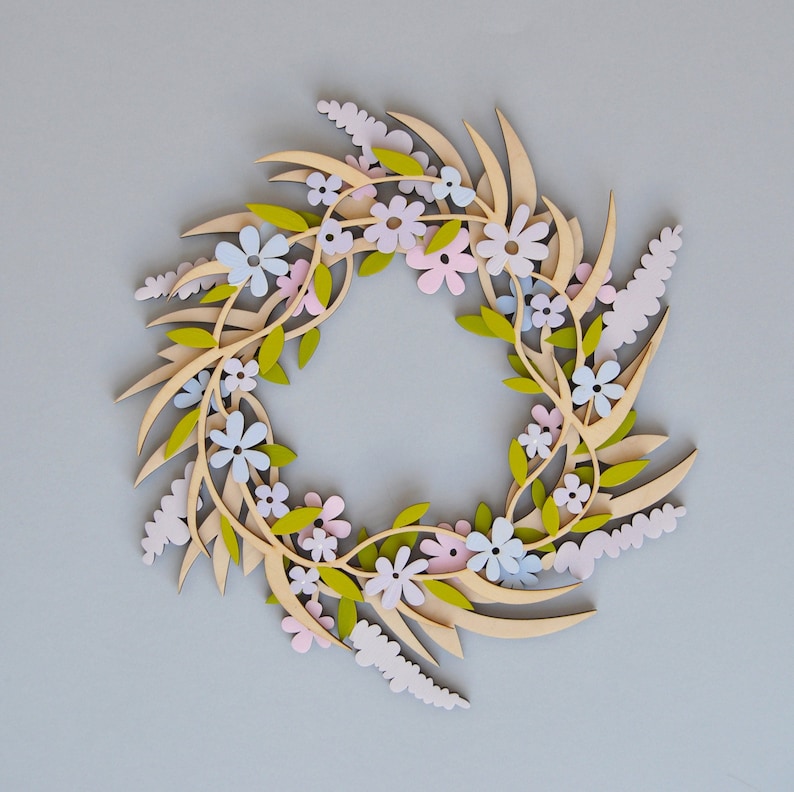 May include: A wooden wreath decorated with paper flowers in shades of pink, blue, and white. The wreath is made of branches and leaves, and the flowers are arranged in a circular pattern.