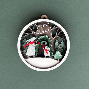 May include: A white wooden ornament with a 3D design of two bunnies decorating a tree with a wreath. The tree is decorated with white lights and the bunnies are wearing red scarves.