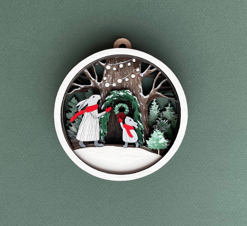 May include: A white wooden ornament with a 3D design of two bunnies decorating a tree with a wreath. The tree is decorated with white lights and the bunnies are wearing red scarves.