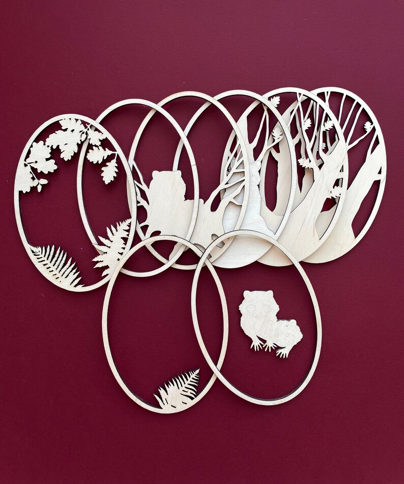 May include: Set of six wooden oval ornaments with various nature-themed cutouts, including trees, ferns, and animals. The ornaments are arranged in a circular pattern, with the cutouts facing outward.