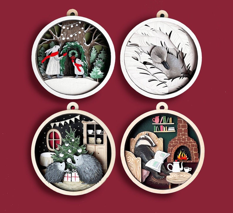 May include: Four round wooden ornaments with different winter scenes. The first ornament features two rabbits in front of a tree with lights. The second ornament features a duck in a nest of leaves. The third ornament features two hedgehogs in front of a Christmas tree. The fourth ornament features a badger sitting in a chair with a book and a fireplace behind it.