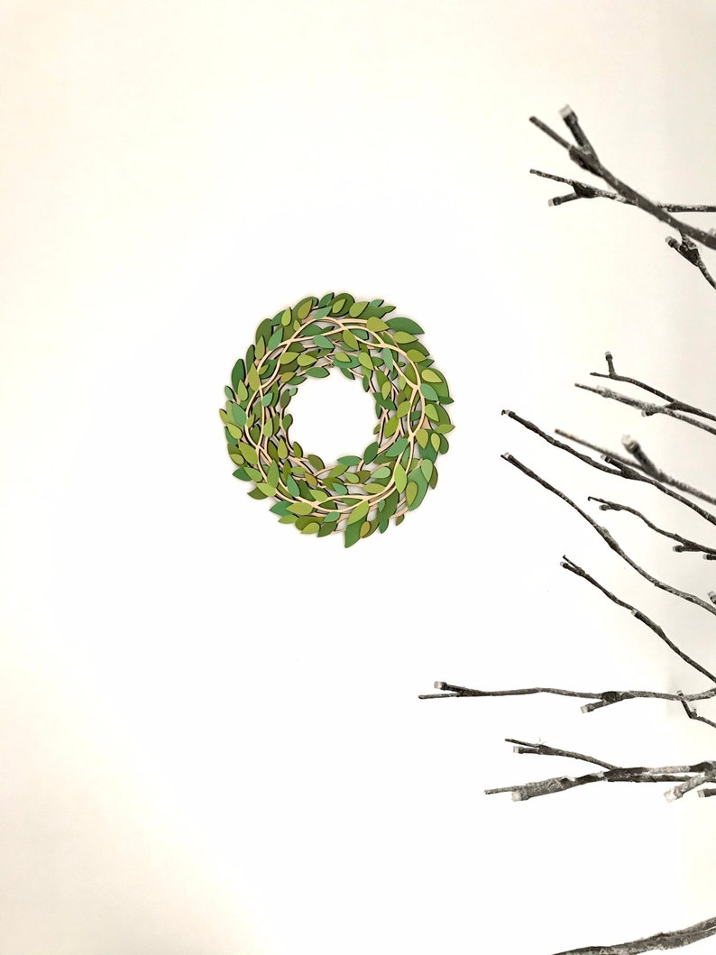 May include: A green leaf wreath with a natural twig base, hanging on a white wall. The wreath is made of individual green leaves and twigs, creating a natural and rustic look.