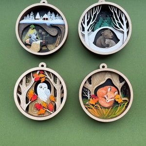 May include: Four round wooden ornaments with intricate paper cutouts depicting winter and autumn scenes. The ornaments feature a mouse in a house, a bear family, an owl in a tree, and a fox sleeping in leaves.