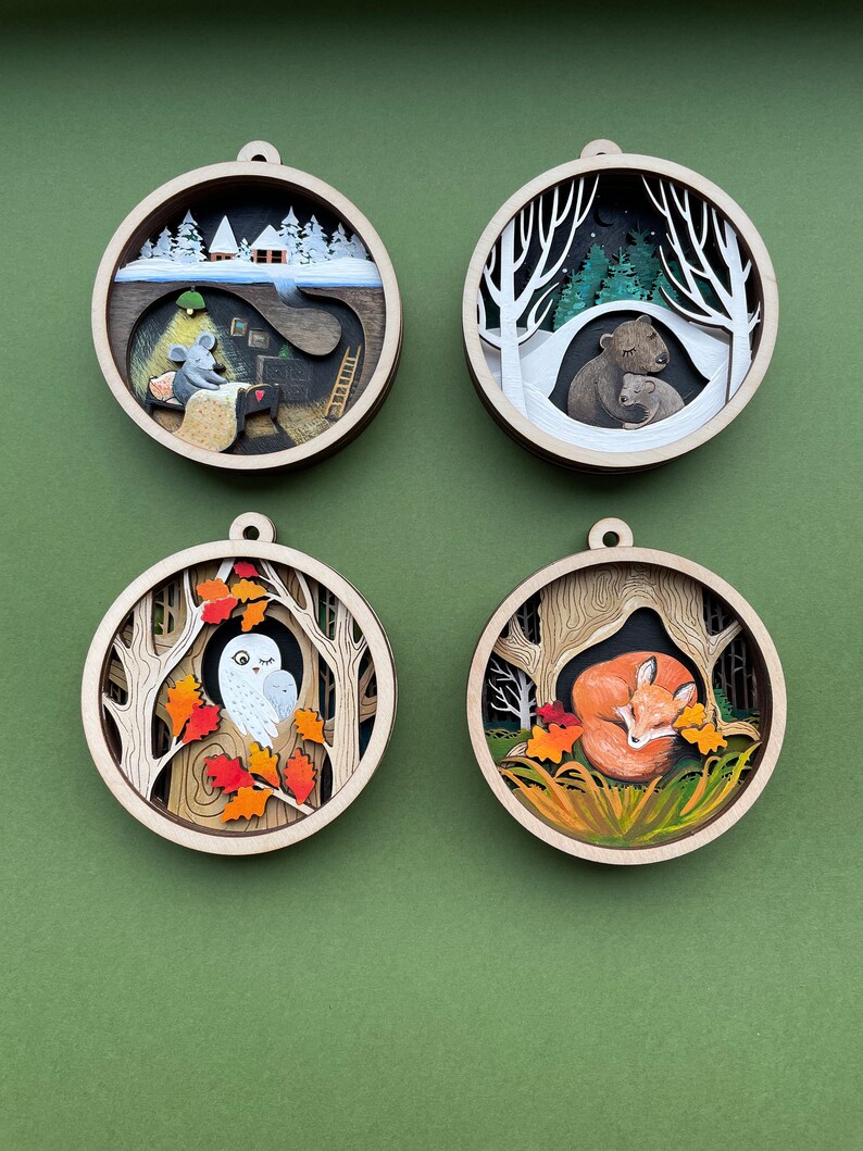 May include: Four round wooden ornaments with intricate paper cutouts depicting winter and autumn scenes. The ornaments feature a mouse in a house, a bear family, an owl in a tree, and a fox sleeping in leaves.