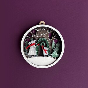 May include: A white wooden ornament with a winter scene. Two rabbits are decorating a tree with a wreath. The ornament has a white background with a brown tree trunk and green leaves. There are white lights strung on the tree.