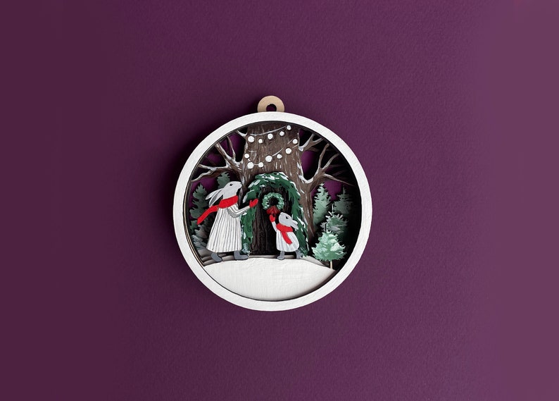 May include: A white wooden ornament with a winter scene. Two rabbits are decorating a tree with a wreath. The ornament has a white background with a brown tree trunk and green leaves. There are white lights strung on the tree.